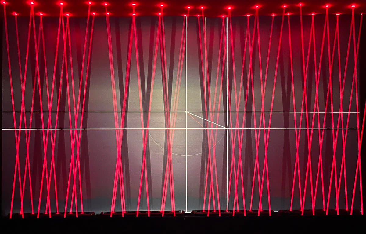 A wide shot of an art installation featuring numerous vertical red laser beams crisscrossing a dark space, intersected by a grid of thin white lines.