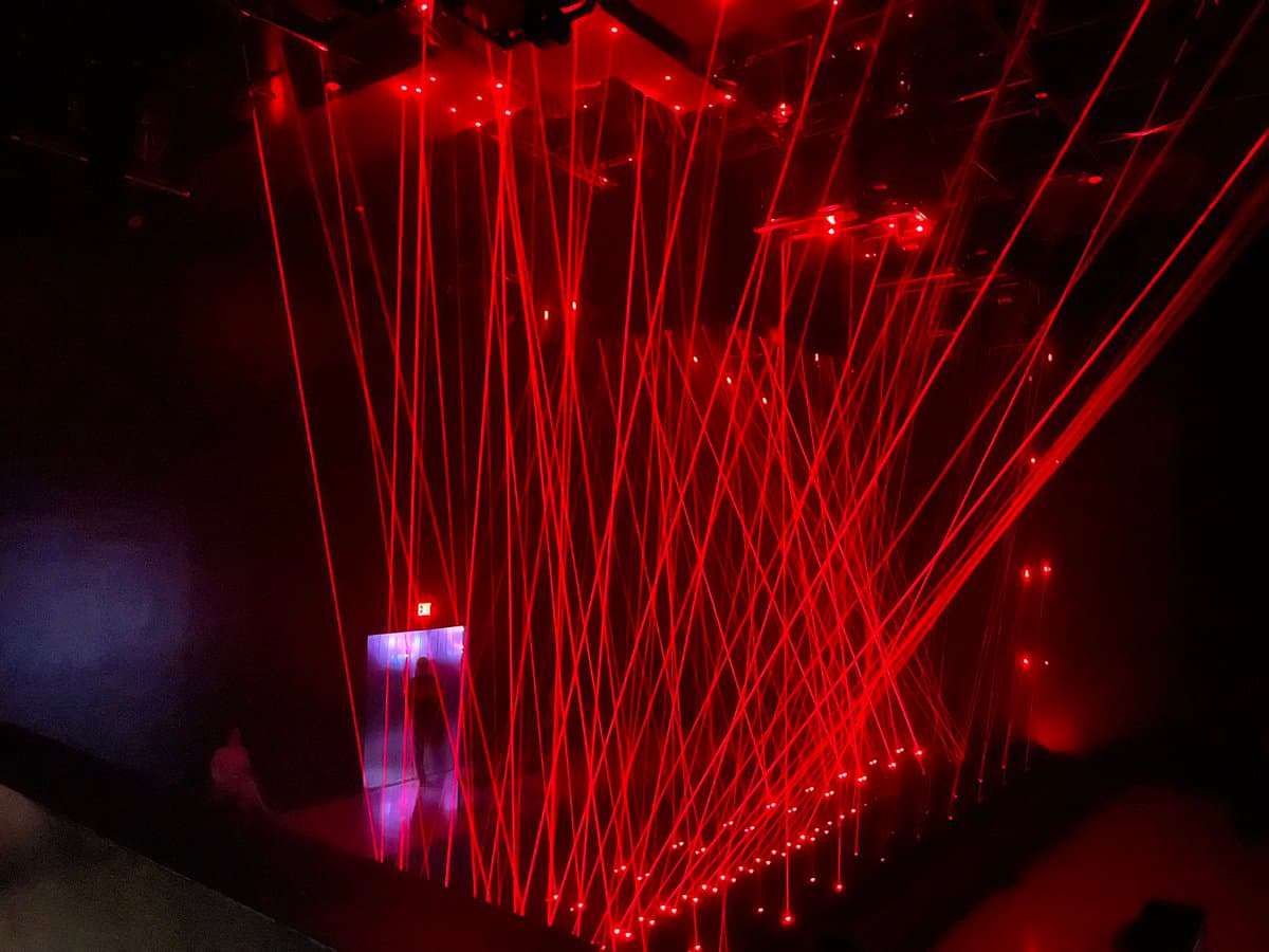 A dark room filled with a dense, crisscrossing web of red laser beams that appear to hang from the ceiling and converge on the floor. In the background, a small doorway is illuminated with blue light, and a red 'EXIT' sign is visible above it.