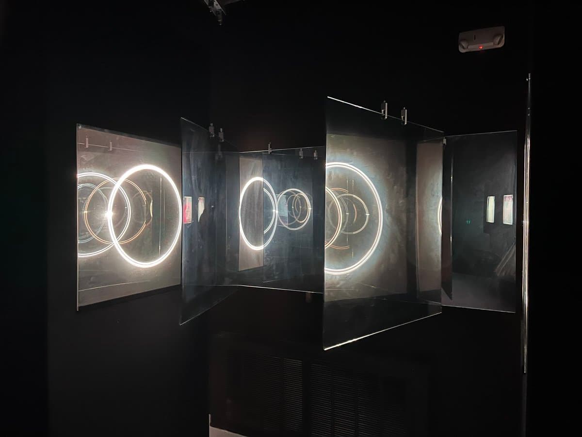 A dark gallery installation featuring multiple angled glass panels displaying glowing, overlapping circular light patterns. The reflective surfaces create a sense of depth and repetition within the dimly lit space.
