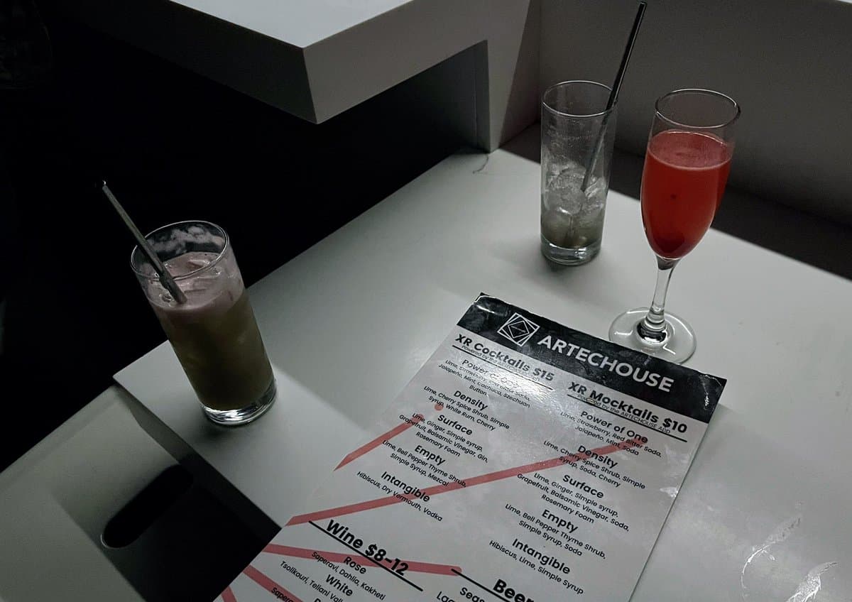 A photo of three mixed drinks and a printed menu on a white table. The menu is for "ARTECHOUSE" and lists "XR Cocktails" and "XR Mocktails." The drinks include a green cocktail in a rocks glass, a clear drink with ice, and a red cocktail in a coupe glass.