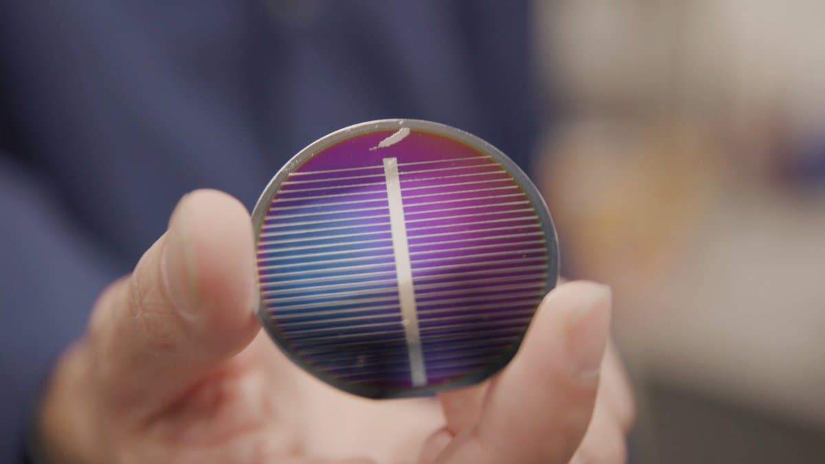 A close-up photograph of a person's hand holding a small, circular solar cell with a purple and blue iridescent surface and thin horizontal lines. The background is blurred, showing a person in a dark blue shirt.