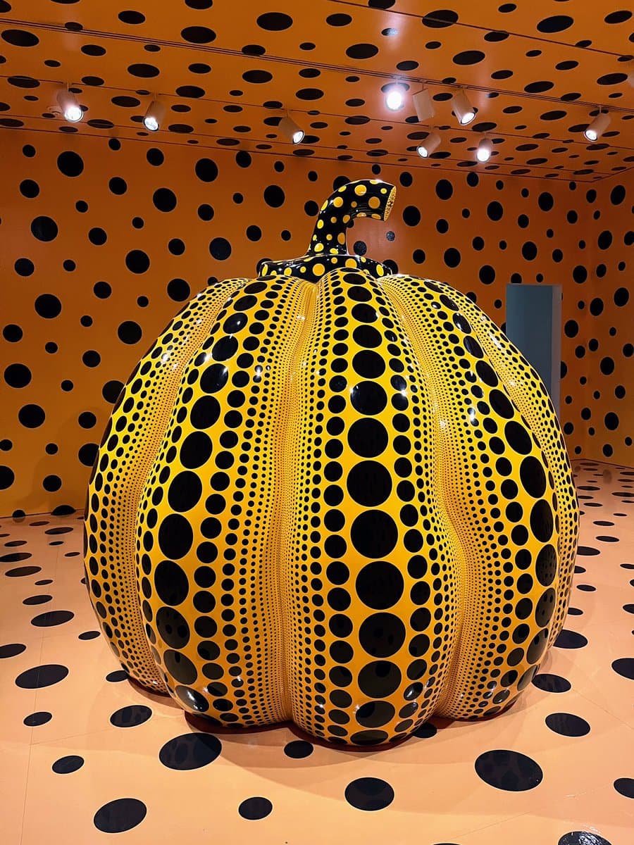 A large, glossy yellow pumpkin sculpture covered in black polka dots sits in the center of a room. The walls, ceiling, and floor are also decorated with the same yellow and black dot pattern, creating an immersive environment. Track lighting is visible on the ceiling.