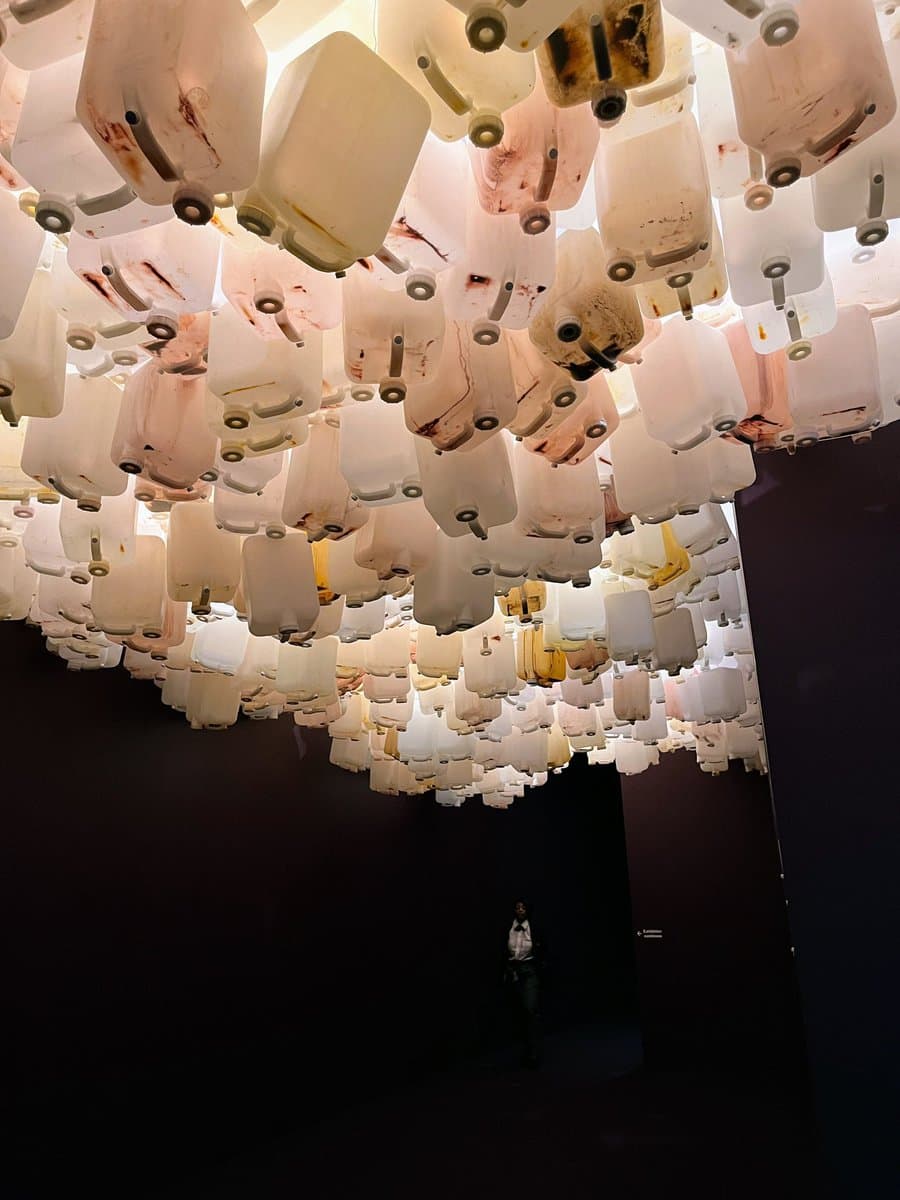 A large-scale art installation featuring hundreds of translucent, stained plastic jugs suspended from the ceiling in a dark gallery space. A single person stands on the floor below, providing a sense of the installation's massive scale.