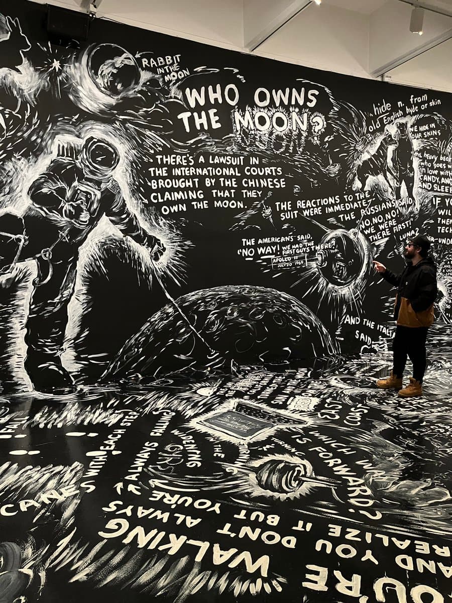 A person stands in a gallery viewing a large, immersive black-and-white mural that covers the walls and floor. The artwork features illustrations of astronauts and the moon, accompanied by extensive text discussing a lawsuit over lunar ownership.