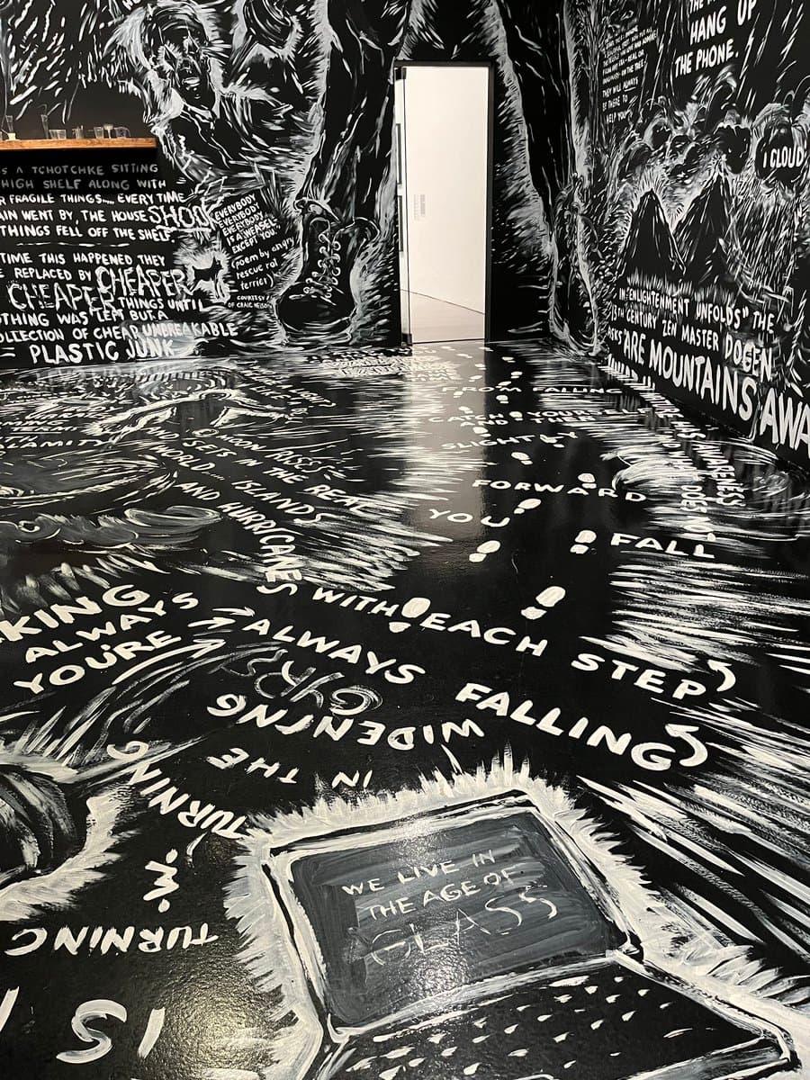 A black-and-white photograph of an immersive art installation featuring a room with walls and floor covered in dense, hand-painted white text and illustrations on a black background. The artwork includes phrases about environmental and social issues, with a central doorway leading to a white space.