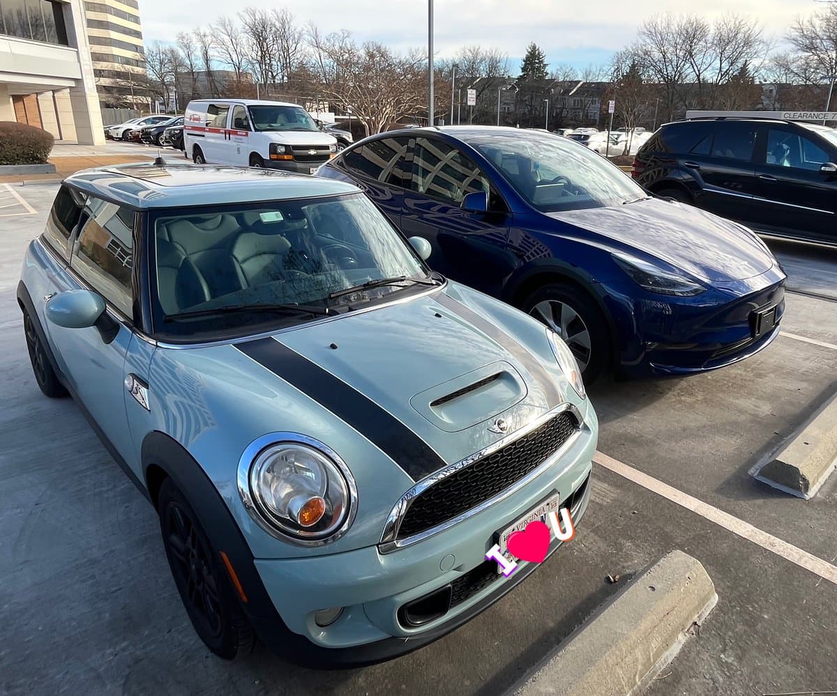 A light blue Mini Cooper with black racing stripes is parked in a parking lot next to a dark blue Tesla Model Y. The Mini's license plate is partially obscured by a digital sticker that reads "I ❤️ U".
