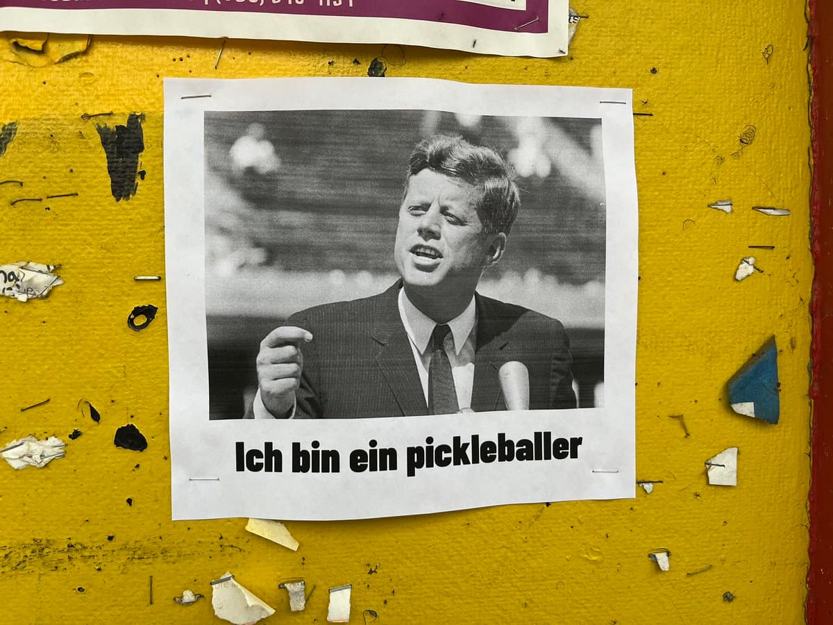 A photograph of a printed poster taped to a weathered yellow wall. The poster features a black-and-white image of a man in a suit pointing forward, with German text below the picture.