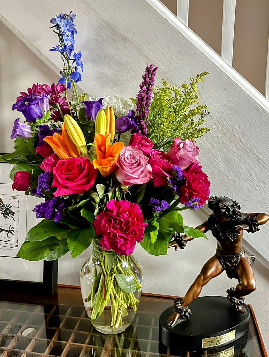 A vibrant bouquet of colorful flowers, including roses, lilies, and carnations, sits in a clear glass vase on a glass table next to a bronze statue of a figure. The background features a white wall and a staircase railing.