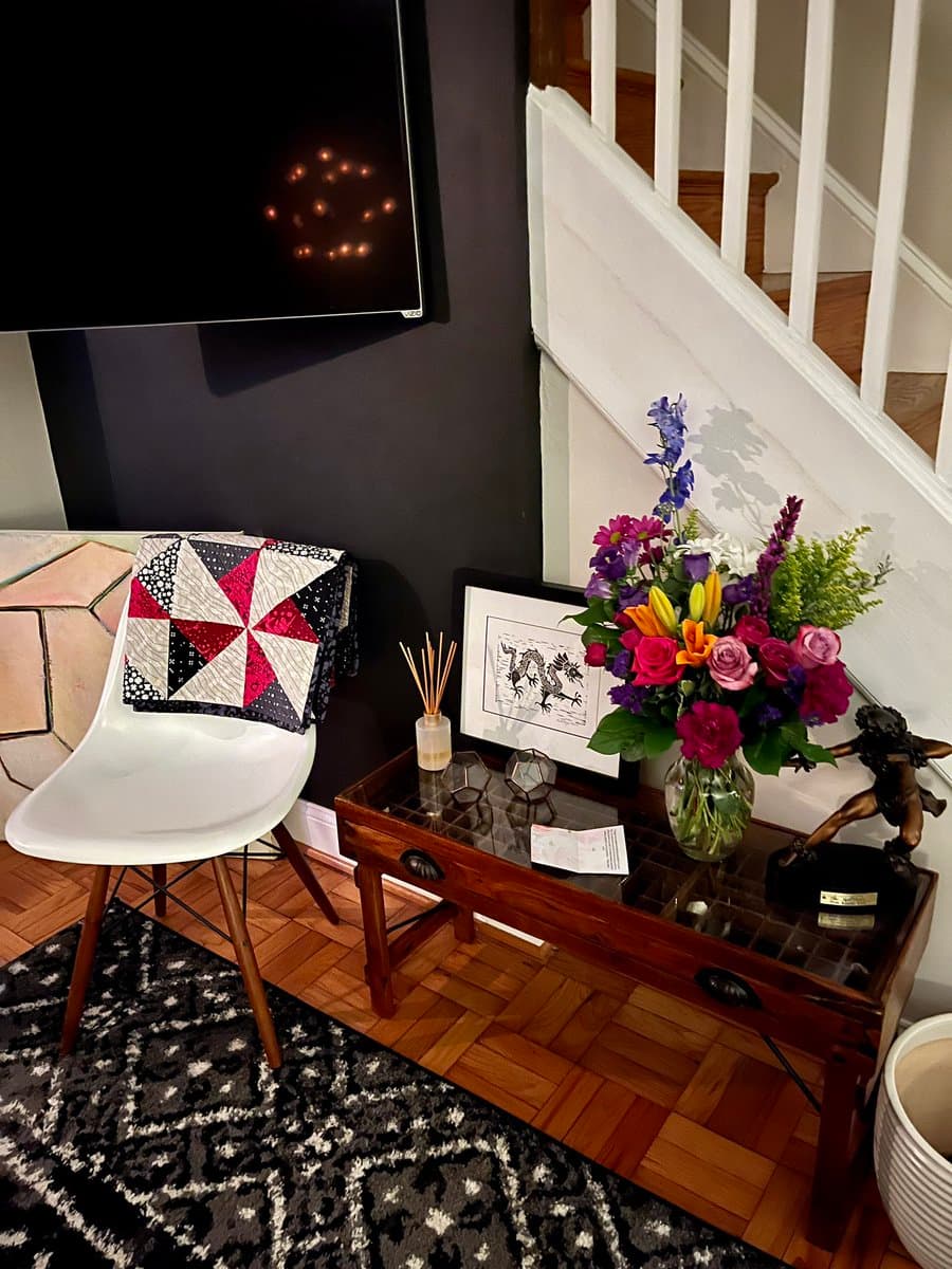 A photograph of a home interior corner featuring a white chair with a red, white, and black quilted pillow, a wooden side table holding a vase of colorful flowers, and a staircase in the background.