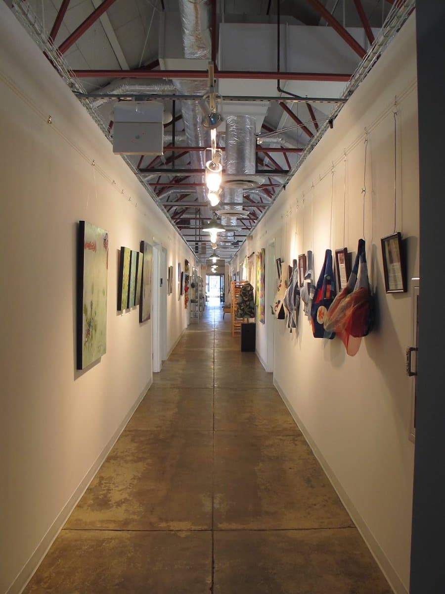 A long, industrial-style hallway with a polished concrete floor and exposed ceiling ductwork, lined with various pieces of artwork and fabric installations hanging from the walls. The space appears to be a gallery or studio area within a converted building.