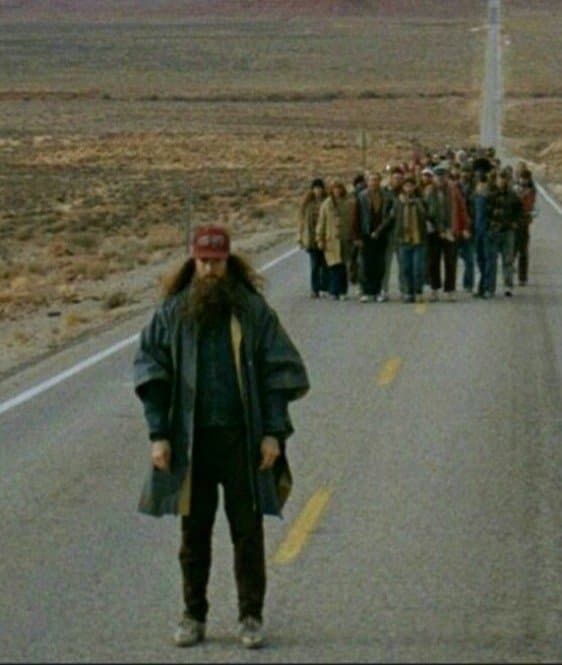 A low-resolution, grainy photograph showing a man with a long beard and a red cap standing in the foreground of a desert highway, with a large group of people walking behind him in a line.