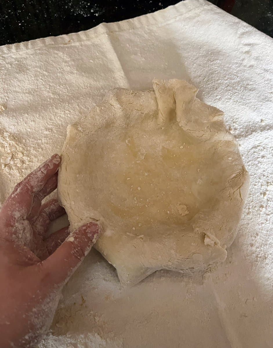 A close-up, top-down view of a person's flour-dusted hands holding a round, uncooked pie crust on a white cloth. The surface is covered in a layer of white flour, highlighting the messy nature of the baking process.