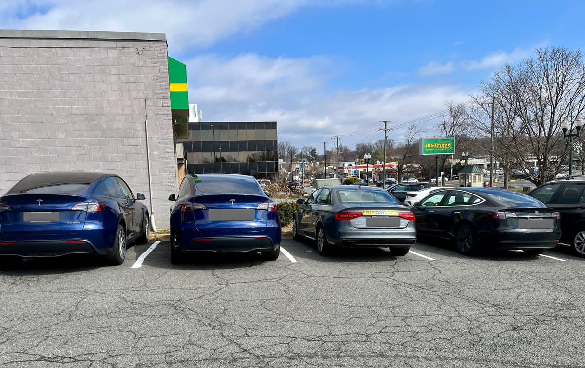 A parking lot with several cars, including multiple blue and black Teslas and a gray Audi, parked in front of a building with a green and yellow sign. In the background, a green sign for "Justyne's" is visible under a partly cloudy sky.
