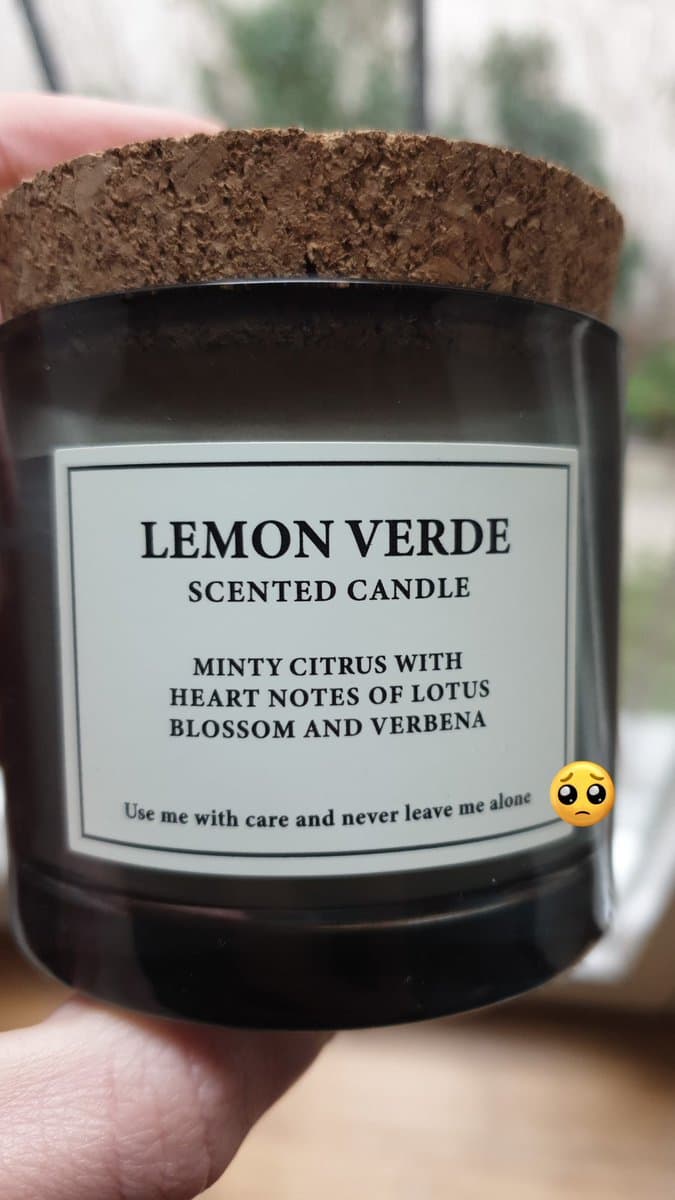 A close-up photograph of a hand holding a dark glass candle jar with a cork lid. The white label on the jar reads "LEMON VERDE SCENTED CANDLE" and includes a humorous warning at the bottom. A pleading face emoji is superimposed on the right side of the label.