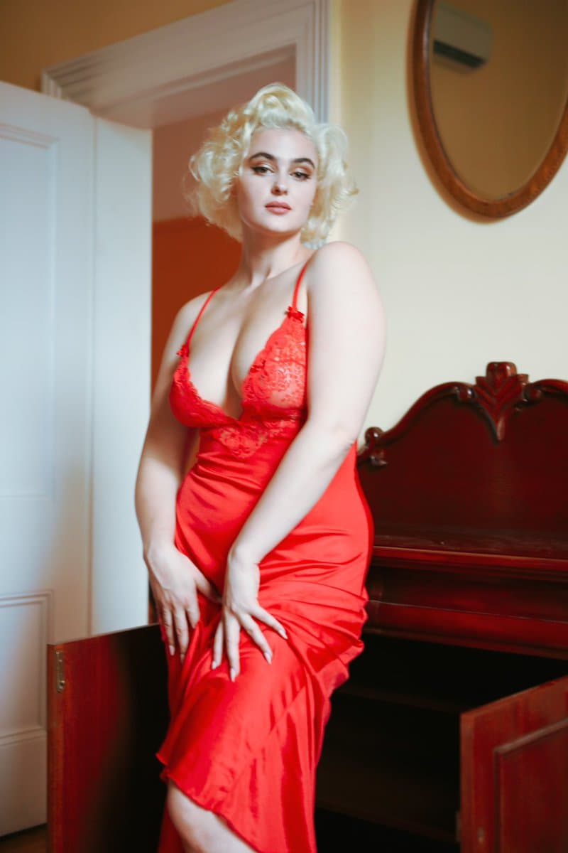 A woman with curly blonde hair stands in a room, wearing a red satin dress with lace detailing on the bodice. She is positioned next to a dark wooden piano, with a white door and a round mirror visible in the background.