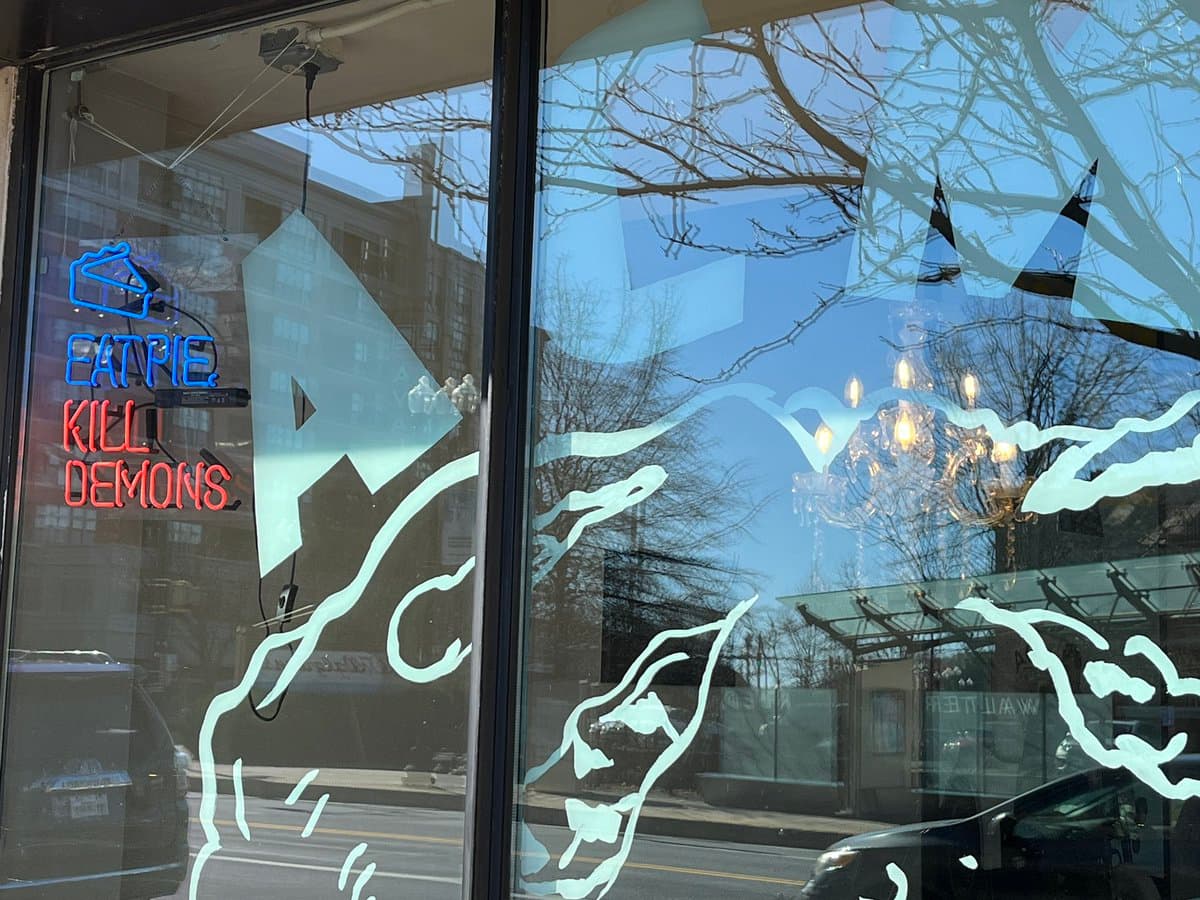 A storefront window featuring a neon sign with a blue pie icon and the text 'EAT PIE' in blue and 'KILL DEMONS' in red, set against a background of white window decals and street reflections.