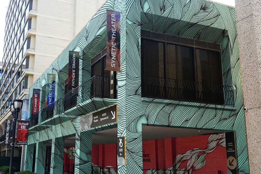 A photograph of the exterior of a building with a distinctive green and white patterned facade, featuring multiple vertical banners and signs. The building is identified as the Synetic Theater, with additional signage for Richard III and a nearby optometrist.