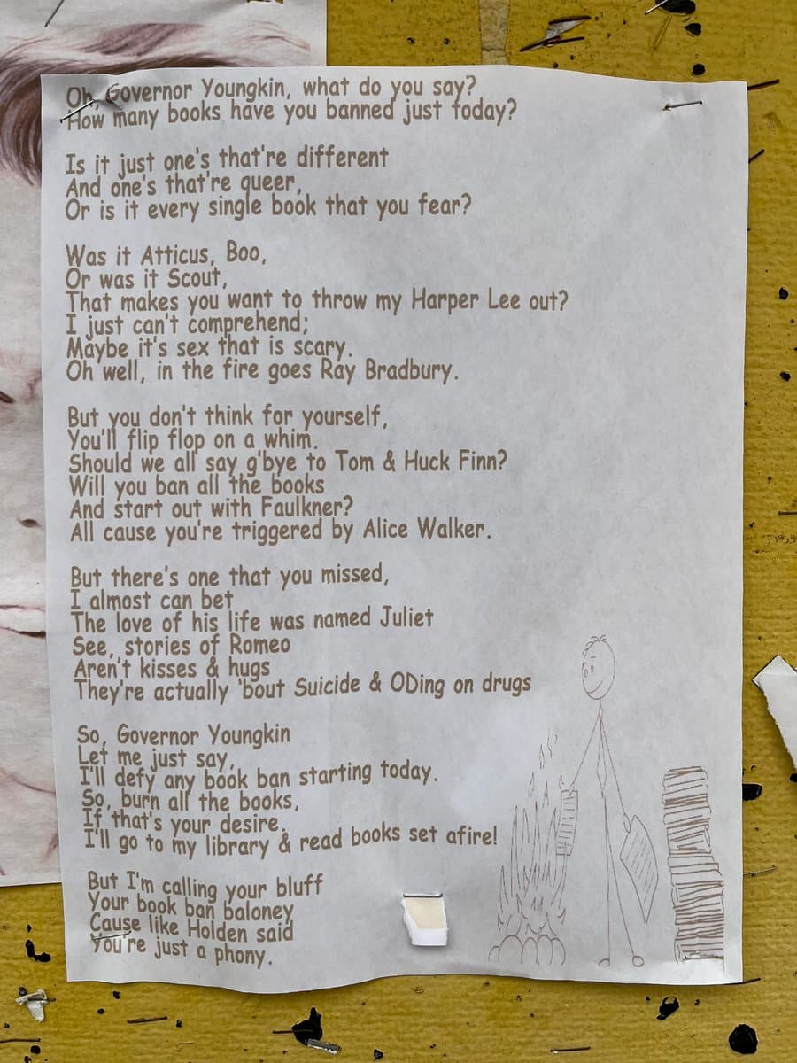 A photograph of a handwritten poem on a sheet of white paper, pinned to a corkboard. The text is a satirical critique of book banning, accompanied by a simple line drawing of a stick figure burning a stack of books.