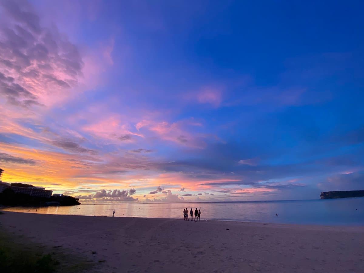 A wide-angle photograph of a beach at sunset, featuring a sky with vibrant pink, purple, and blue clouds. Silhouettes of several people stand on the sand near the water's edge, with a resort building visible on the left and a small island on the right.