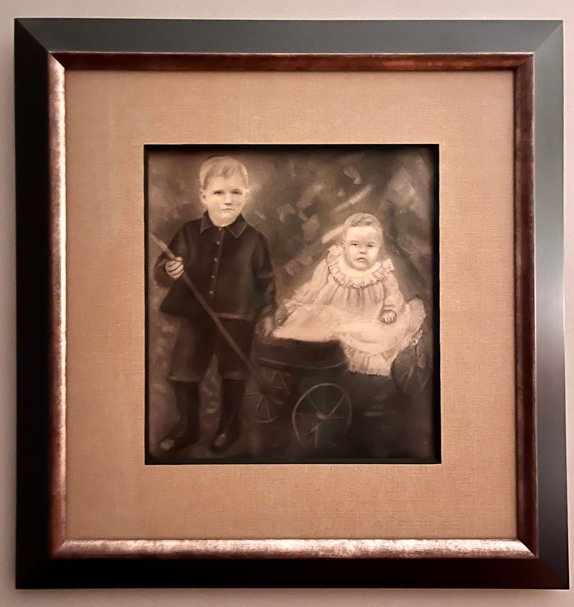 A framed, sepia-toned painting depicting a young boy standing next to a baby sitting in a pram. The boy holds a long stick, and the baby wears a light-colored dress with a ruffled collar. The artwork is mounted in a wide, tan-colored mat within a dark, beveled frame.