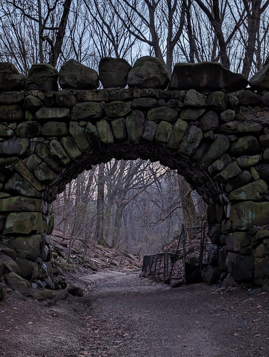 A stone archway constructed from large, moss-covered rocks spans a dirt path in a wooded area. Bare trees and a metal gate are visible through the archway, suggesting a late autumn or winter setting.