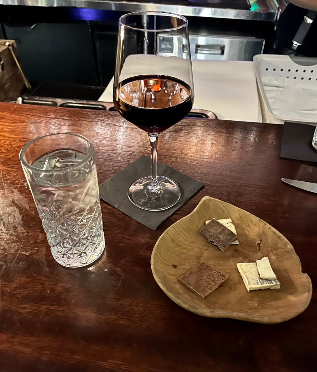 A glass of red wine, a glass of water, and a wooden plate with pieces of dark and white chocolate sit on a wooden bar counter.