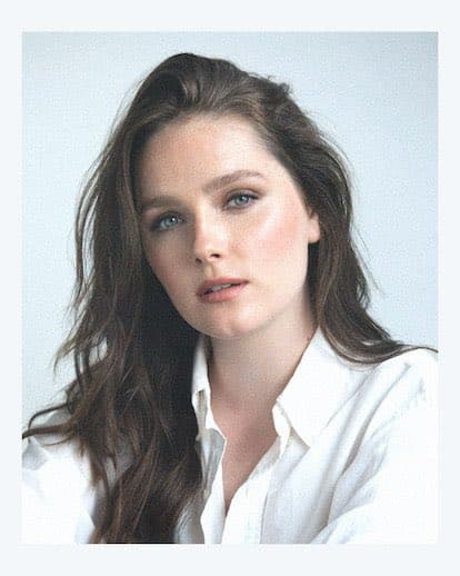 A portrait of a woman with long, wavy brown hair and blue eyes, wearing a white collared shirt against a plain light background. She is looking directly at the camera with a neutral expression.