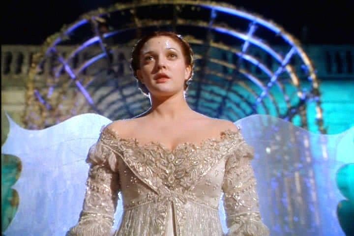 A woman with dark hair and a decorative headpiece stands in front of a large, illuminated circular structure, wearing an ornate, off-the-shoulder silver gown with long sleeves. She has large, translucent white wings attached to her back, suggesting a fairy or angelic character in a theatrical or cinematic setting.