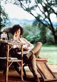 A woman with curly hair sits at a small outdoor table, resting her chin on her hand while looking toward the camera. She is wearing a light-colored shirt and dark pants, seated on a wooden chair in a garden setting with trees and greenery in the background.