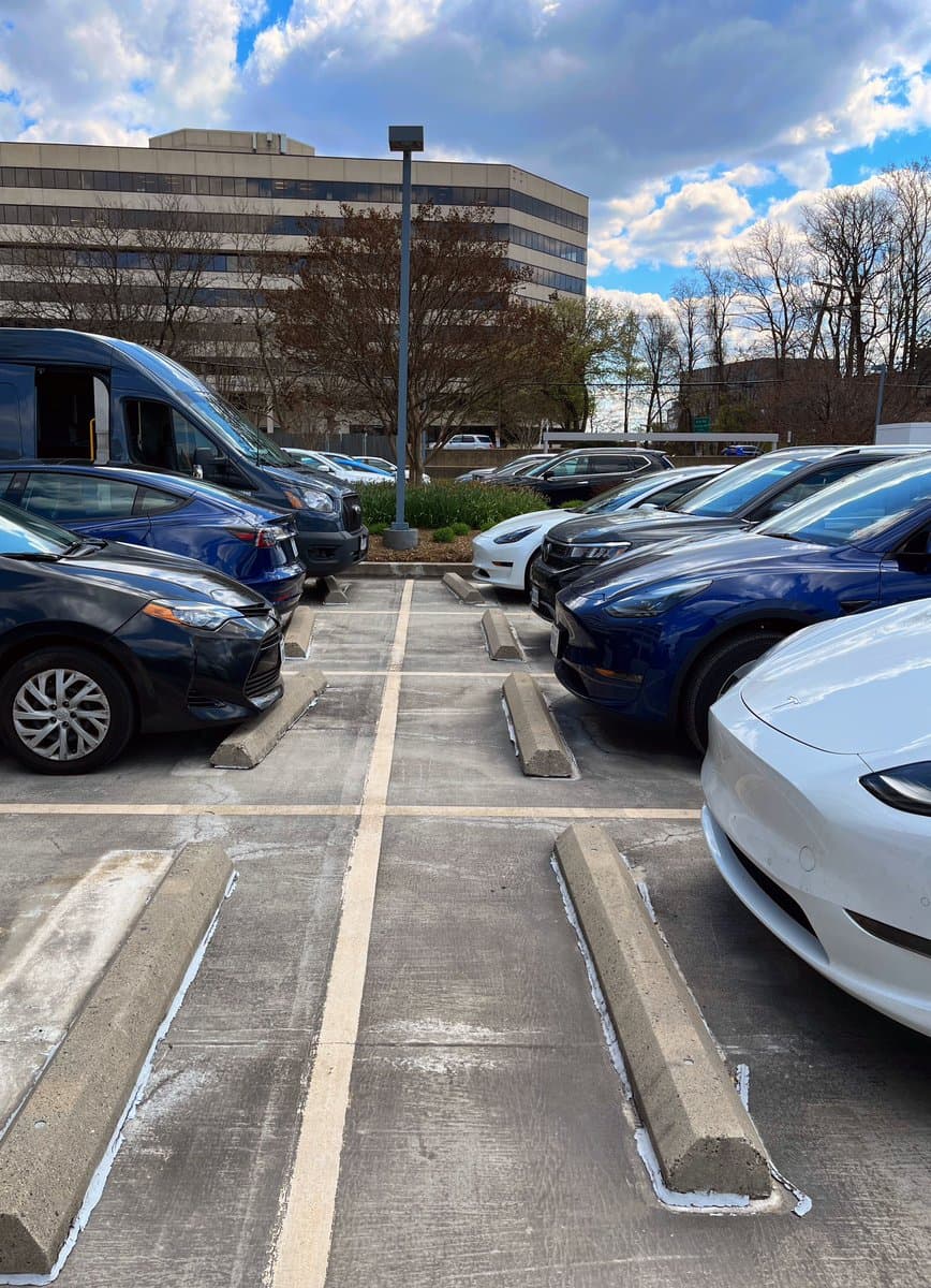 A parking lot filled with numerous blue and white Tesla vehicles parked in rows, with a large office building and trees in the background under a partly cloudy sky. The image is a photograph of a parking lot scene.