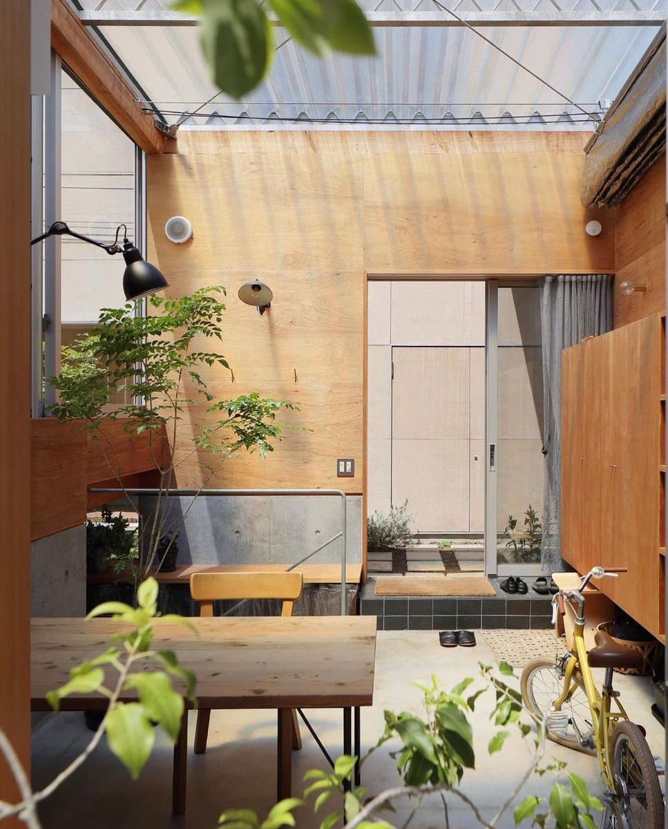 A photograph of a modern, minimalist interior space featuring light wood paneling, a concrete floor, and a glass roof. The room contains a wooden table and chair, a potted plant, a black floor lamp, and a yellow bicycle parked near a wooden cabinet.