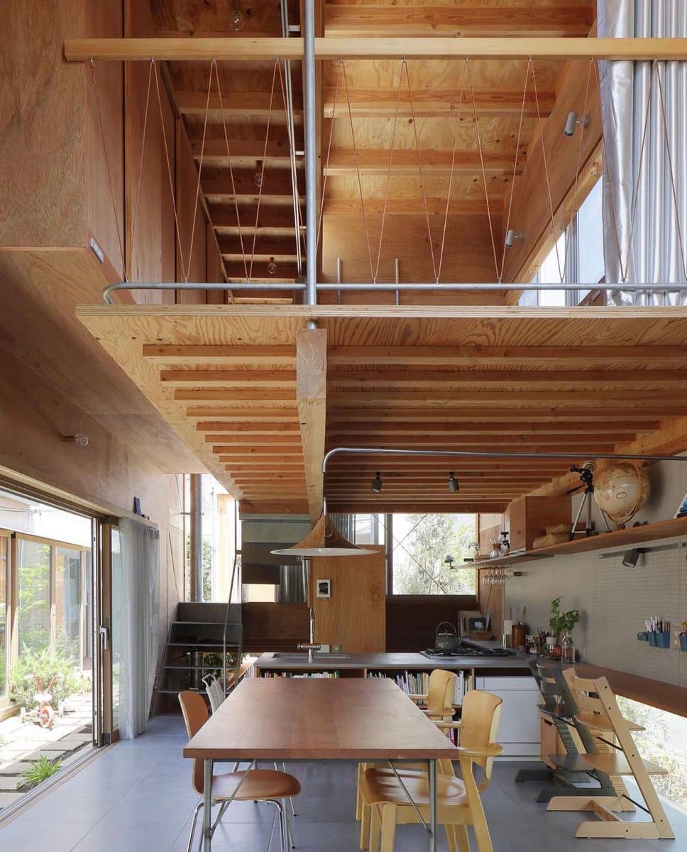 A photograph of a modern, open-plan interior featuring extensive use of light wood, including a long dining table, exposed ceiling beams, and a loft area with a suspended staircase. The space includes a kitchen area, large windows, and various household items, creating a bright and airy atmosphere.