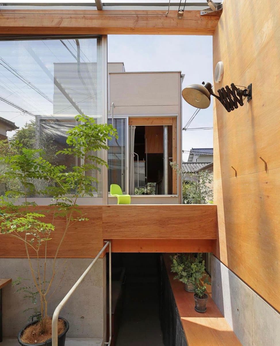 A photograph of a modern architectural space featuring exposed wooden beams, a glass window, and a small balcony with a green chair. A potted tree stands in the foreground, and an adjustable wall lamp is mounted on the wooden wall to the right.