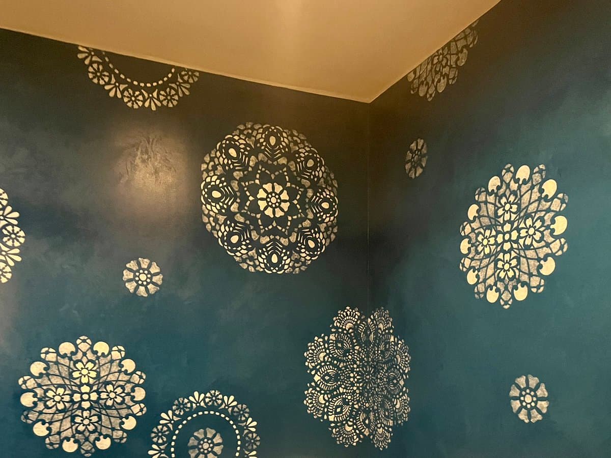 A corner of a room with dark teal walls decorated with large, intricate white mandala and floral stencils. The ceiling is painted a light cream color, and the lighting creates a soft, moody atmosphere.