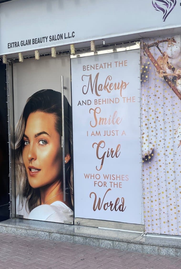 A photograph of a storefront window display for a beauty salon, featuring a large portrait of a woman and a sign with a humorous quote.