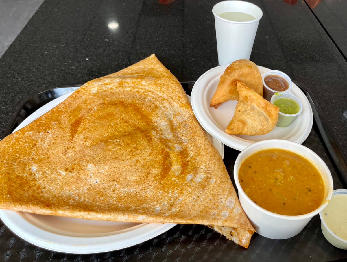 A tray of South Indian food featuring a large folded dosa on a white plate, a bowl of sambar, a plate with two samosas and small cups of chutney, and a cup of water. The meal is arranged on a black tray on a dark countertop.