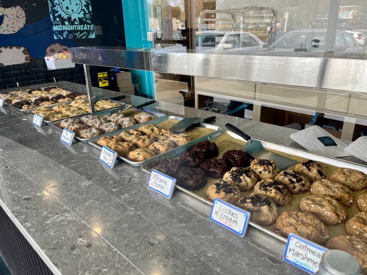 A display case at a bakery filled with trays of various baked goods, including cookies and brownies, with handwritten labels in front of each tray.