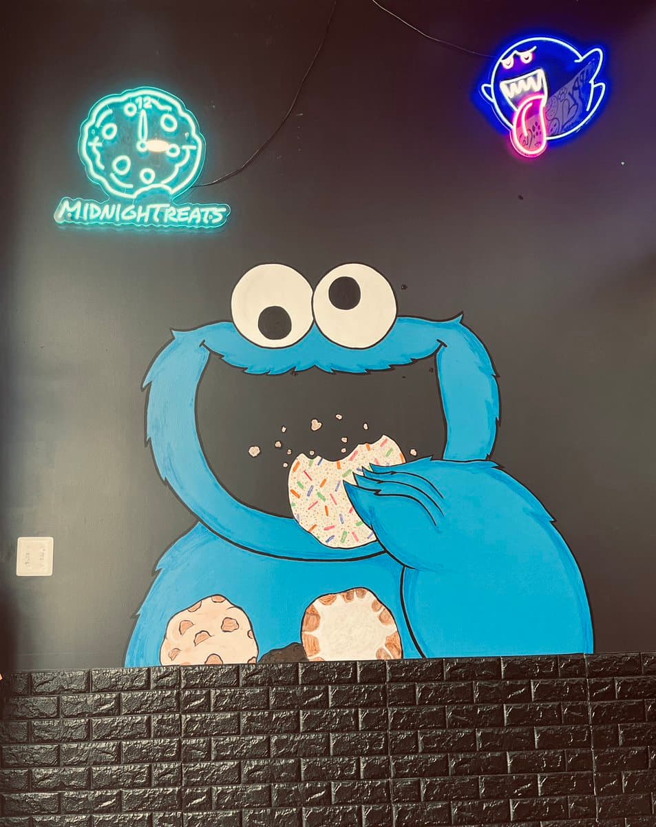 A wall mural of a blue, Cookie Monster-like character eating a sprinkle-covered cookie, set against a dark wall with a faux-brick base. Above the character are two neon signs: a teal clock reading 'MIDNIGHT TREATS' and a blue ghost with a pink tongue.