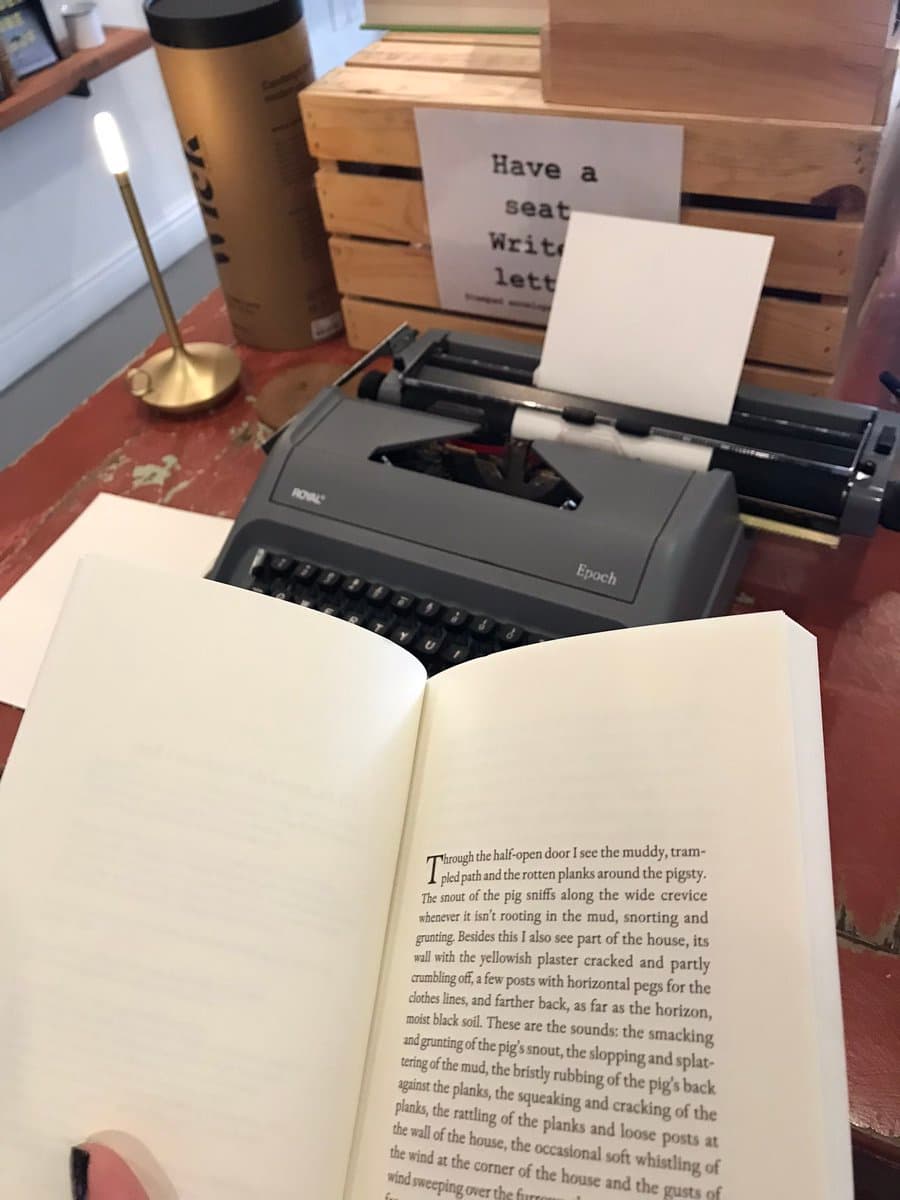 A first-person view of an open book resting on a wooden table in a bookstore, with a vintage Royal Epoch typewriter and a sign in the background. The scene includes a small brass lamp and a cardboard coffee cup.