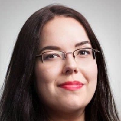 A headshot portrait of a woman with long dark hair, wearing thin-framed glasses and red lipstick, smiling gently against a plain light gray background. This appears to be a professional profile photo.