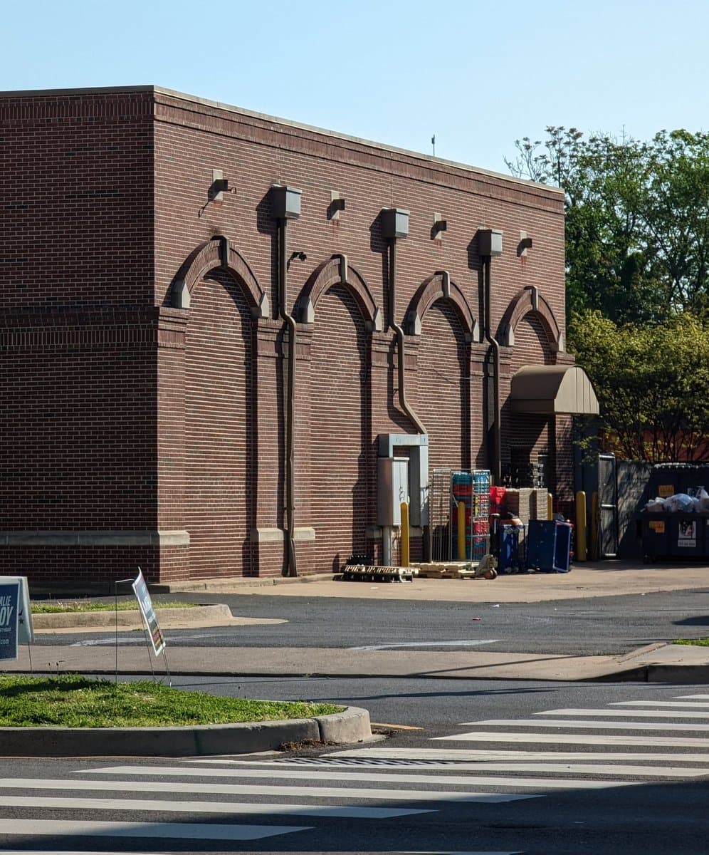 A photograph of a red brick building featuring a row of decorative arched recesses and external drainage pipes, with a paved parking lot and crosswalk in the foreground.