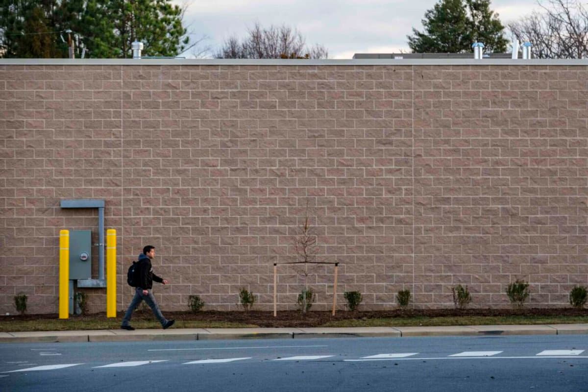 A photograph showing a person walking past a long, blank stone wall of a building, likely a pharmacy, with a small tree and utility box visible in the foreground.
