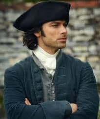 A man with dark hair and a beard is shown from the chest up, wearing a dark blue 18th-century style coat, a white cravat, and a black tricorn hat. He has his arms crossed and is looking off to the side against a blurred stone wall background. The image appears to be a still from a period drama or film.