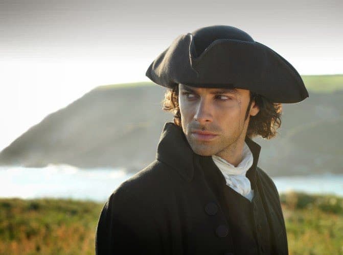 A man with wavy brown hair wearing a black tricorn hat and a dark coat with a white cravat, standing outdoors with a coastal landscape in the background.