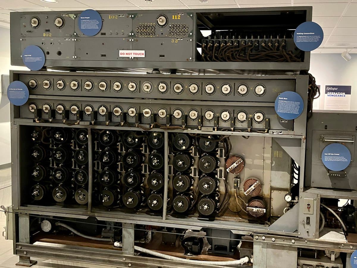 A large, vintage piece of electronic equipment, likely a WWII-era computing or communications machine, displayed in a museum setting. The machine features multiple rows of dials, switches, and vacuum tubes, with several blue informational placards attached to its surface. A sign on the upper left panel reads 'DO NOT TOUCH.'