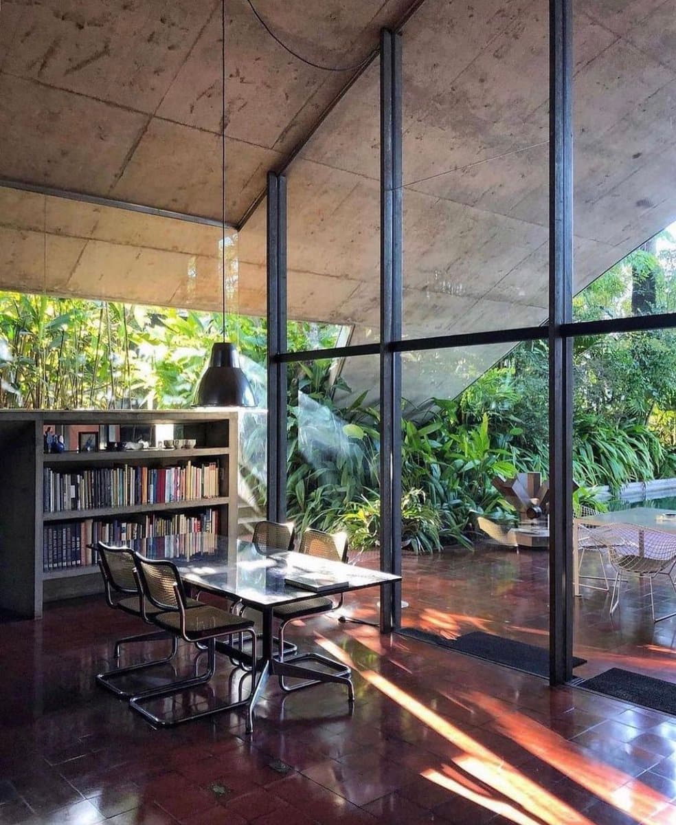 A photograph of a modern interior space featuring a glass-walled room with a dining table, chairs, and a bookshelf, surrounded by lush green foliage outside.