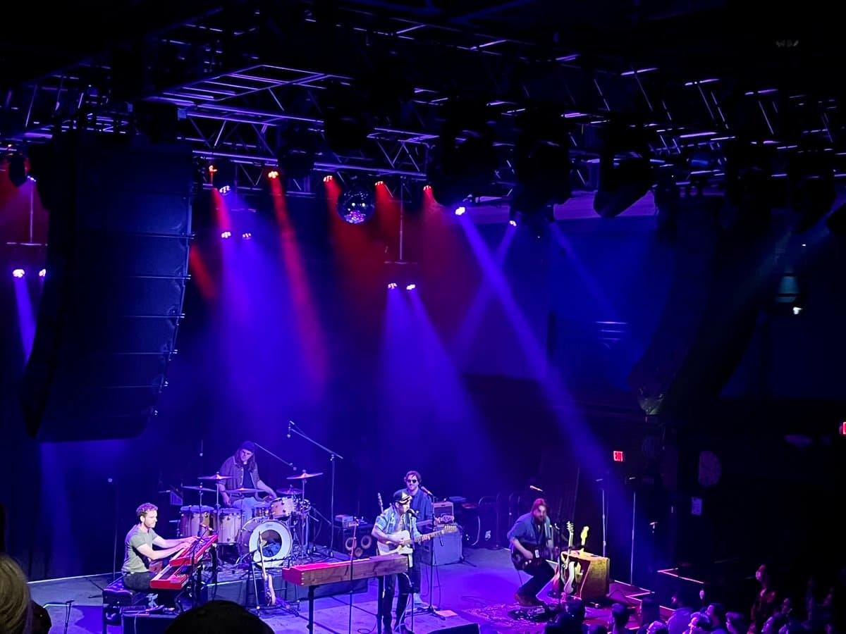 A wide shot of a band performing on a concert stage under dramatic purple and red stage lighting. The image captures the musicians, including a drummer, keyboardist, and guitarists, along with the overhead lighting rig and a disco ball.