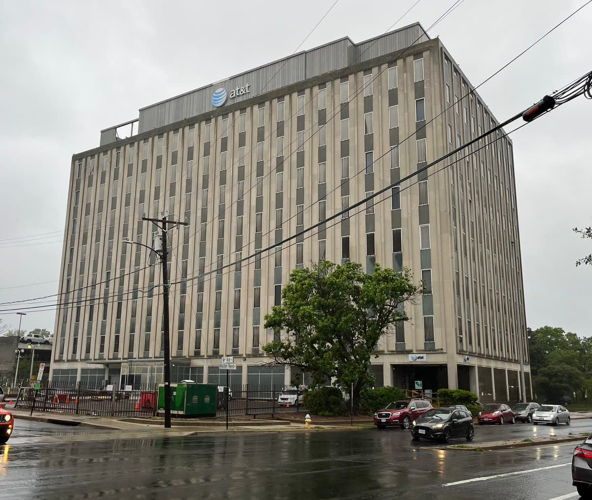 A large, multi-story office building with an AT&T logo on its roof, photographed from across a wet street on an overcast day. Several cars are parked or driving in the foreground, and a street sign for "Walter Reed" is visible near the sidewalk.
