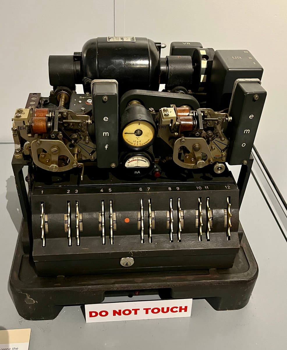 A photograph of a vintage black electromechanical telegraph or teleprinter machine displayed on a table, featuring various dials, levers, and a row of numbered switches. A white sign with red text reading 'DO NOT TOUCH' is placed in front of the device.