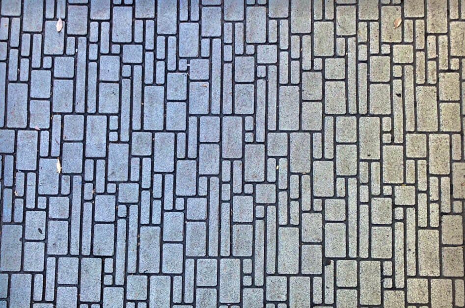 A top-down view of a paved surface featuring a geometric pattern of rectangular and square stones in varying shades of gray and beige. The image captures the texture and arrangement of the tiling, with a few small leaves scattered across the surface.